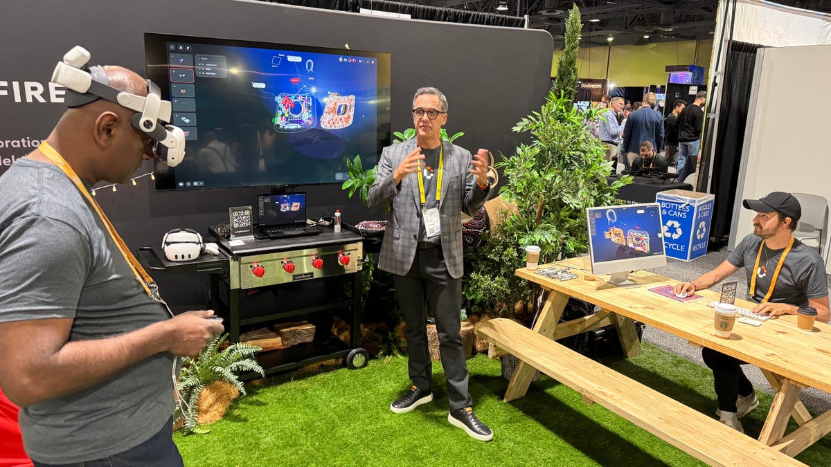 A man in a VR headset and another man sitting at a bench using an iMac work on the same 3D model, displayed on a TV between them.